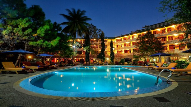Una piscina con sillas de descanso y sombrillas, rodeada de palmeras y un edificio de varios pisos con ventanas iluminadas.