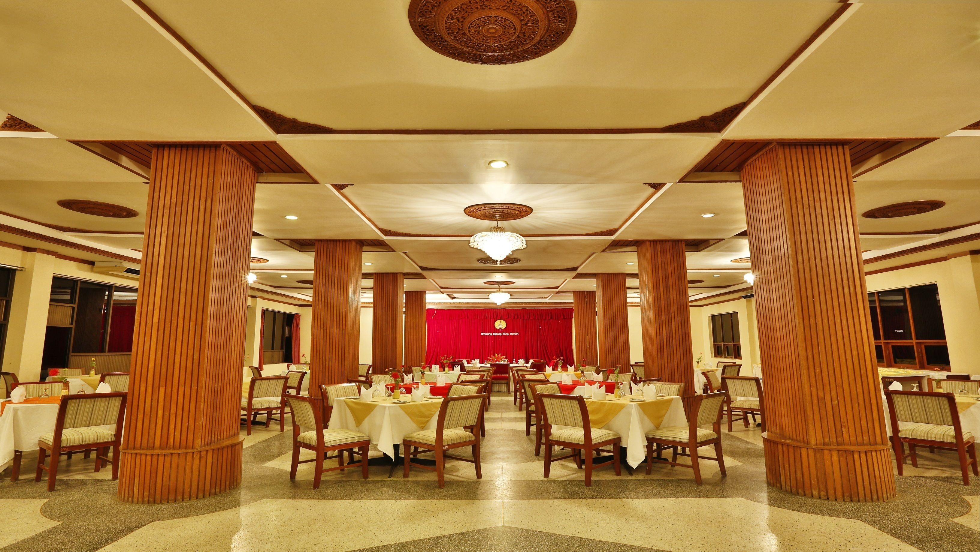 Une salle de banquet avec des tables rondes, des colonnes en bois et un rideau rouge en arrière-plan.