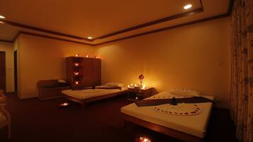 Two beds with white bedspreads and rose-petal-adorned pillows, a wardrobe, and a small table with a vase of flowers.