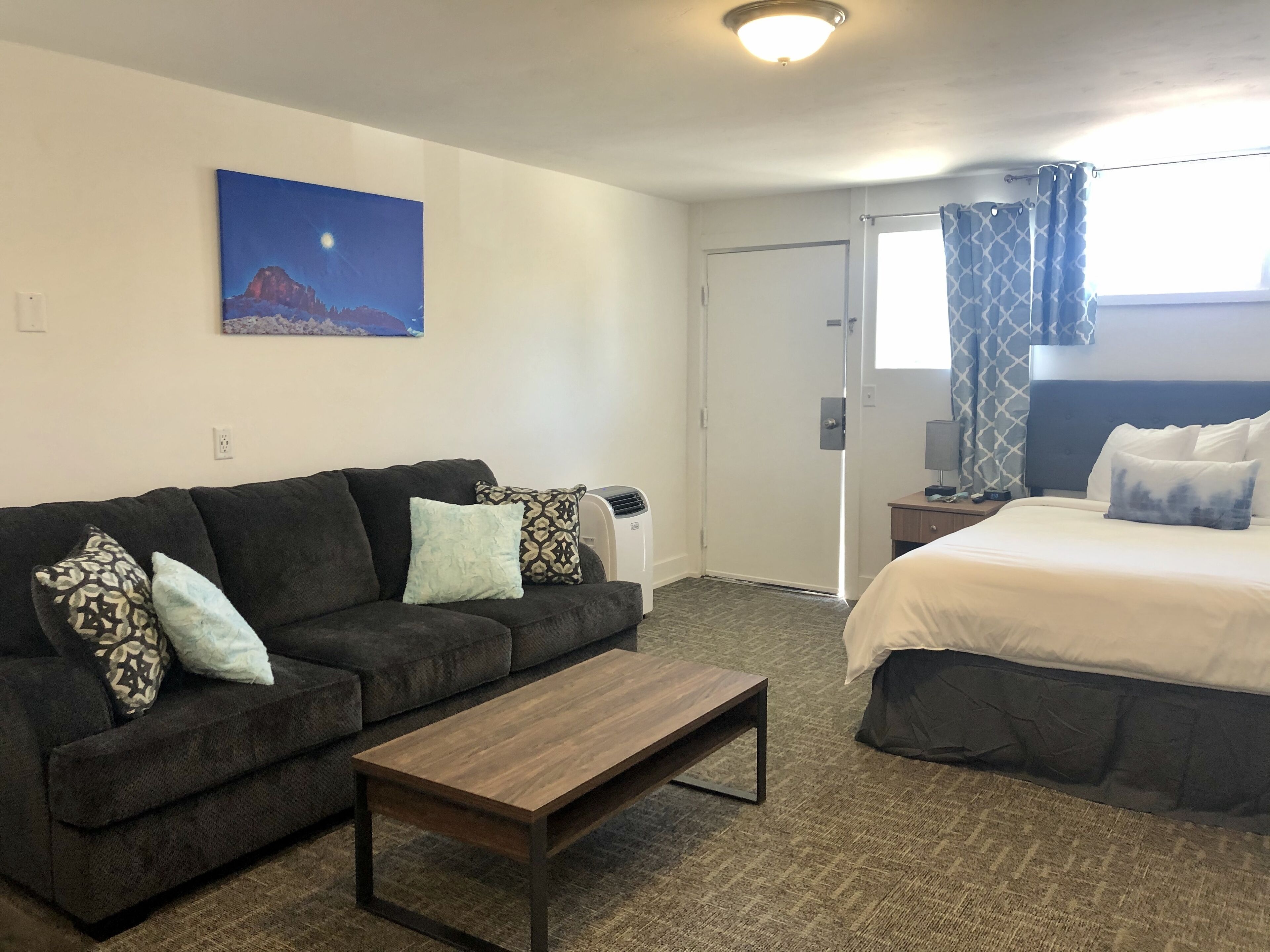 A hotel room with a dark-colored sofa, a wooden coffee table, a bed with white bedding, a nightstand with a lamp, a painting on the wall, and a window with patterned curtains.