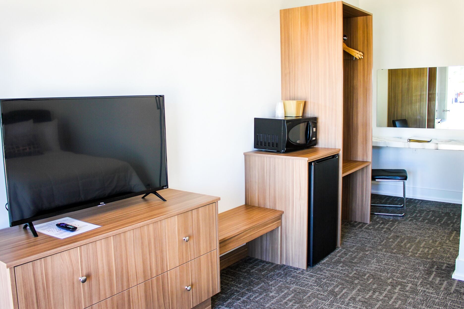 Habitación de hotel compacta con cama, televisión, microondas, refrigerador pequeño, cómoda de madera y espejo.
