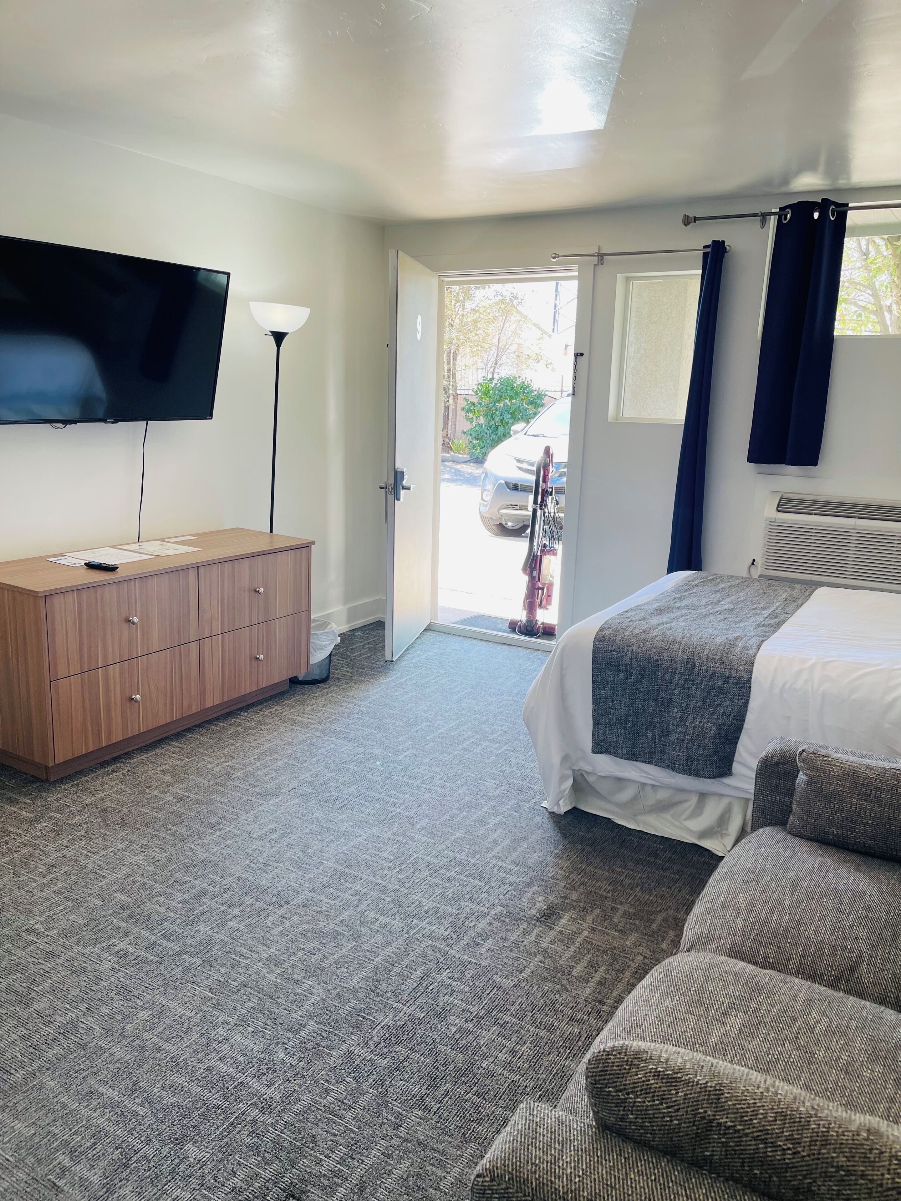 A bedroom with a bed, a TV mounted on the wall, a wooden dresser, and a door leading outside.