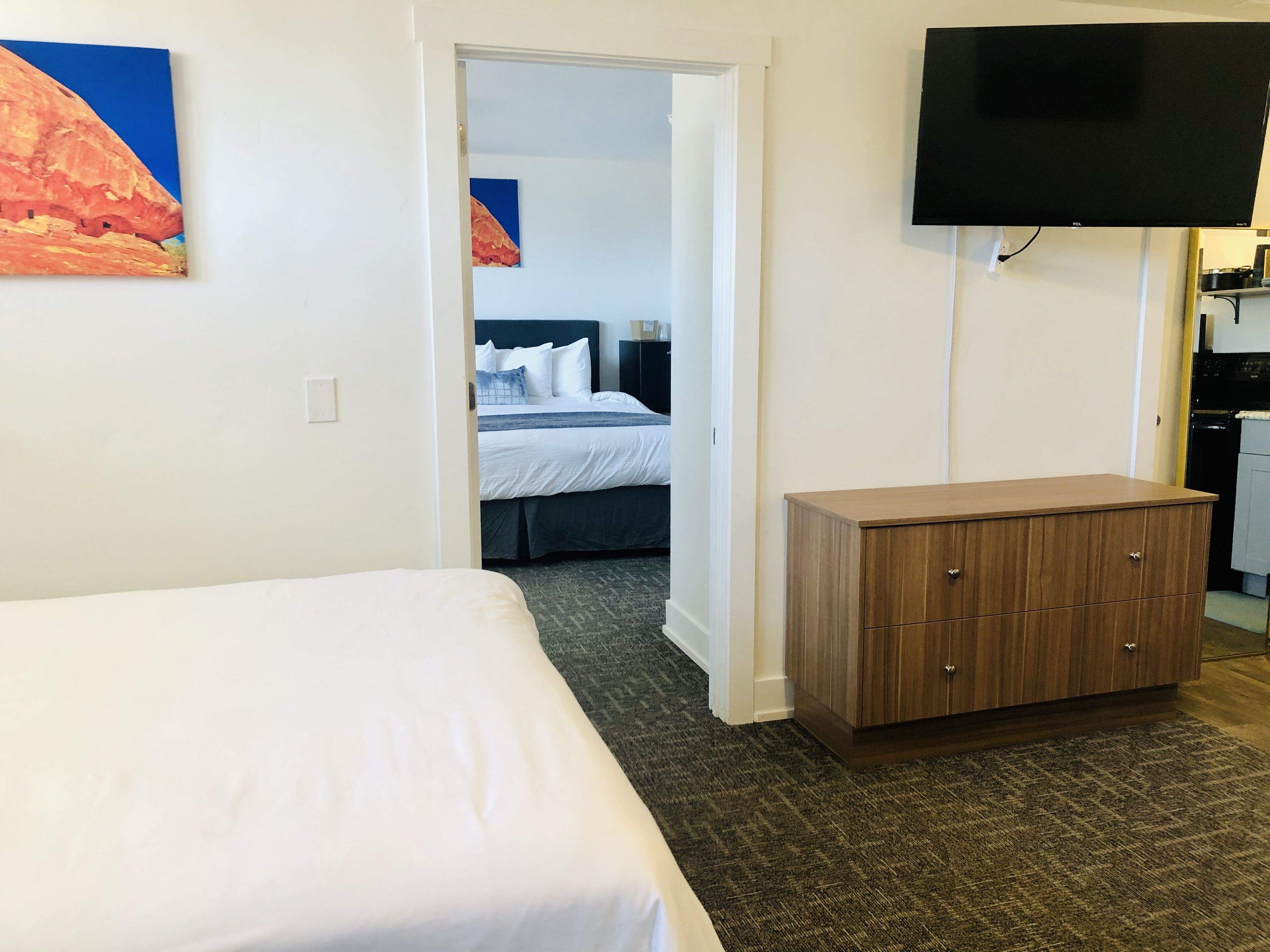 A hotel room with a bed, a wooden dresser, a flat-screen TV, and a door leading to another room.