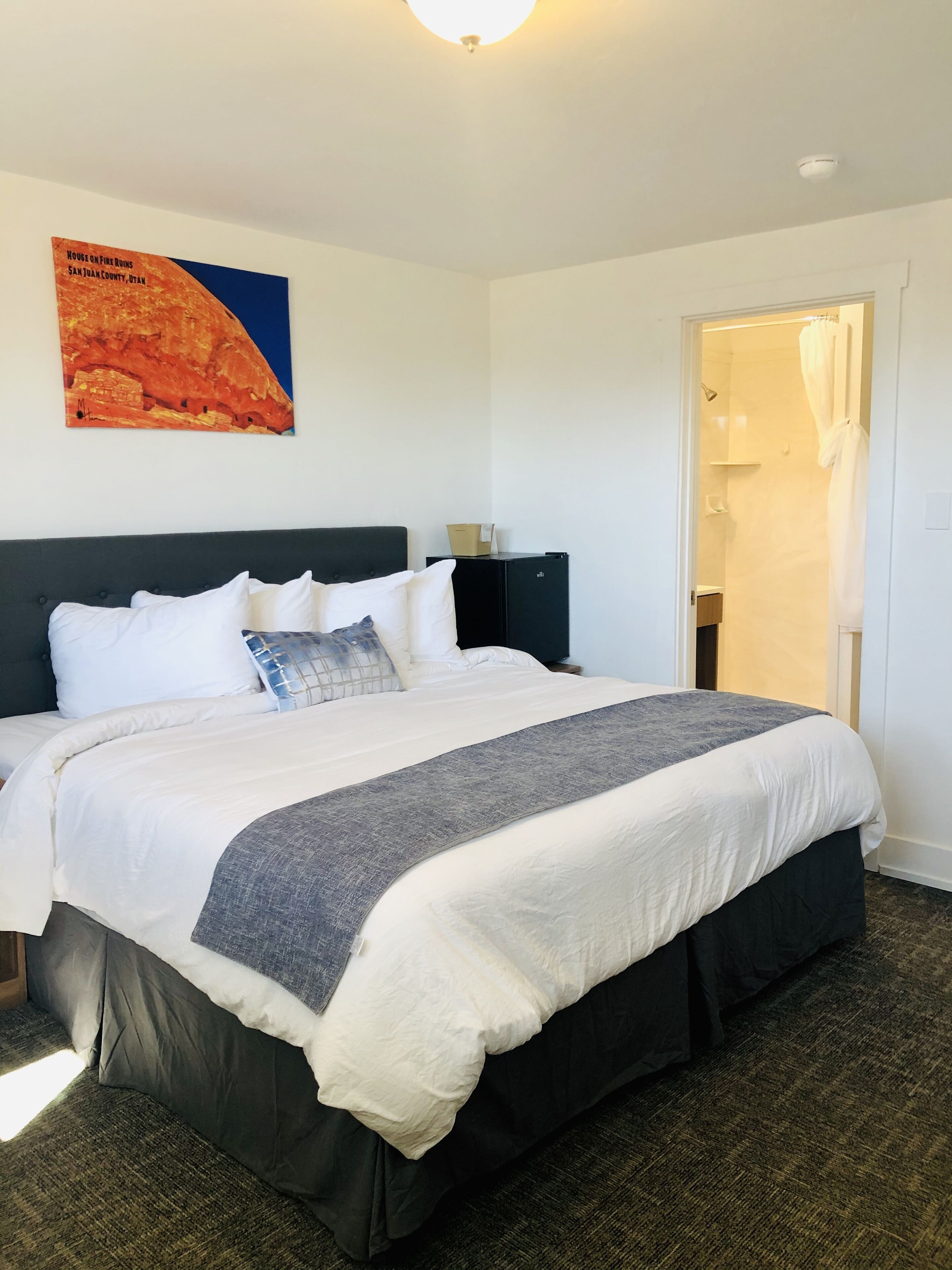 A neatly made bed with white and gray bedding, a dark headboard, and a painting of a red rock formation on the wall.