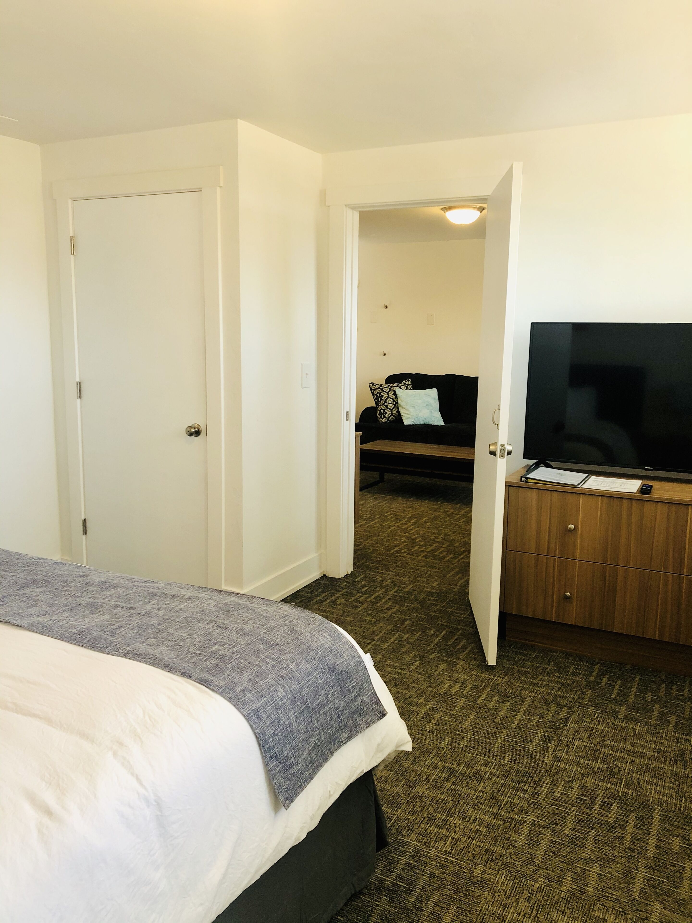 A hotel room with a bed, a TV, a desk, and a door leading to another room.