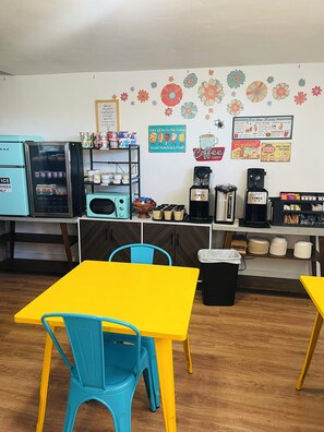A café with a yellow table and blue chairs, a vintage refrigerator, a coffee machine, and a menu board on the wall.