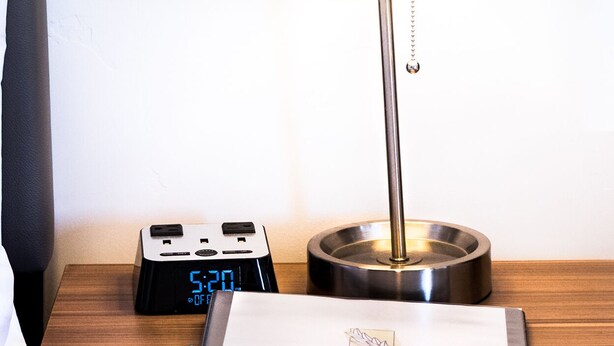 Una mesita de noche de madera con un reloj digital, una lámpara y una tarjeta llave de hotel.