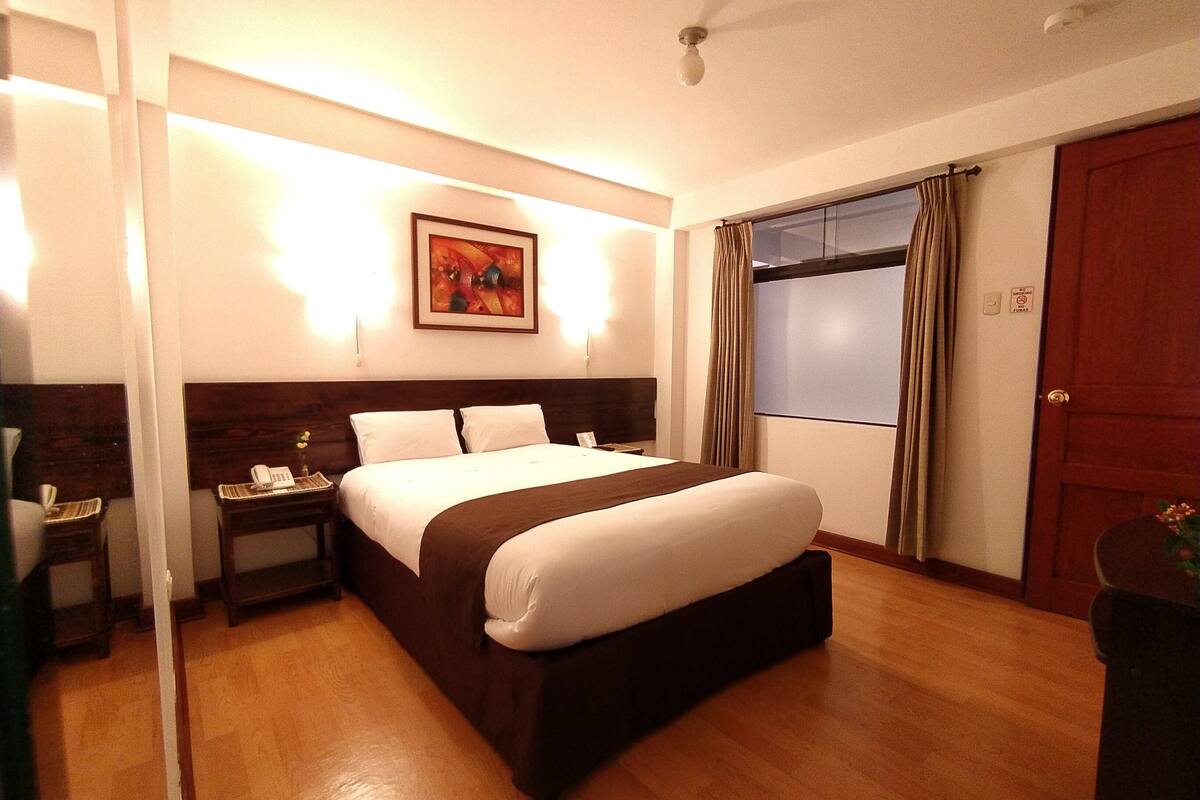 A hotel room with a large bed, a bedside table with a lamp, a painting on the wall, and a window with curtains.