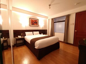 A hotel room with a large bed, a bedside table with a lamp, a painting on the wall, and a window with curtains.