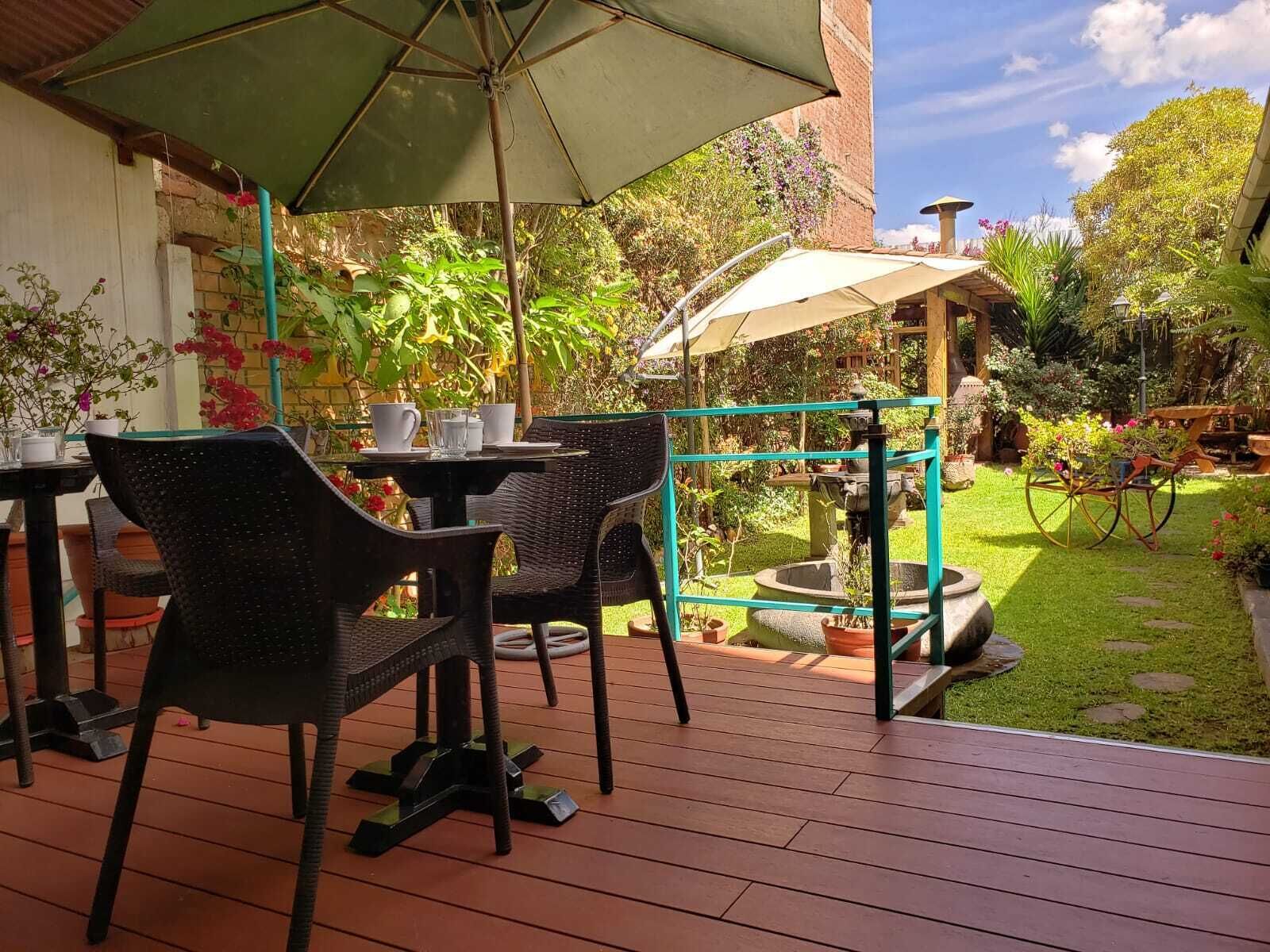 An outdoor patio area with wicker chairs and tables, a green umbrella, and a small garden with a fountain.