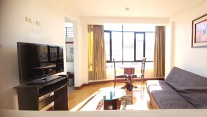A living room with a flat-screen TV, a sofa, a dining table with chairs, and a large window with curtains. - Illas Inn (Cusco)