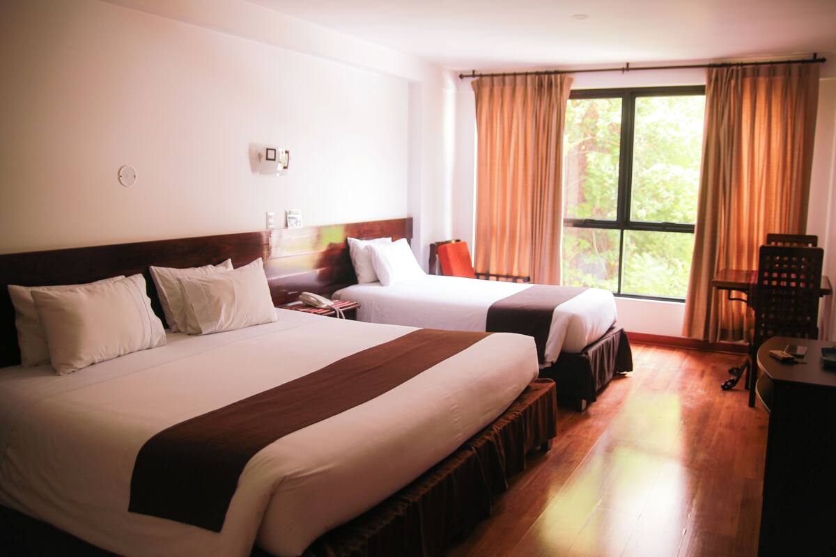 A hotel room with two beds, a wooden headboard, a nightstand, a chair, and a window with curtains.