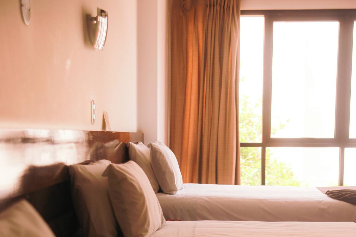 A hotel room with two beds, a window with curtains, and a wall clock.