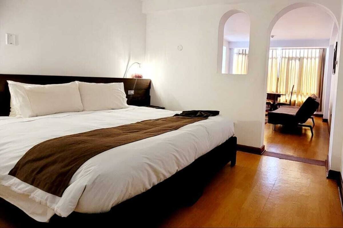 A hotel room with a large bed, a nightstand, and a desk with a chair. There is a window with curtains and a view of the outside.