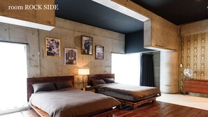 Ein Zimmer mit zwei Betten, Holzboden, einer Betonwand mit gerahmten Bildern, einem Holzkasten und einem Fenster mit Blick.
