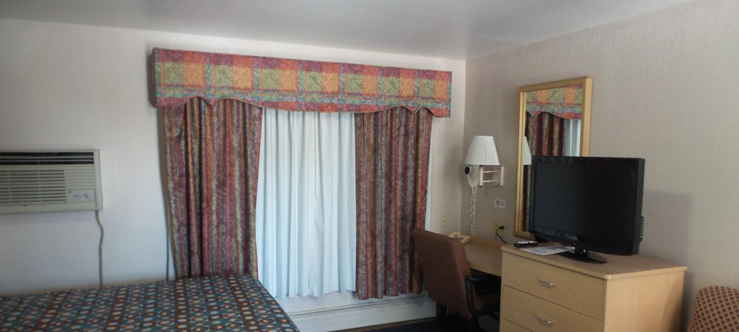 Habitación de hotel con cama, escritorio con computadora, televisor, espejo y teléfono.