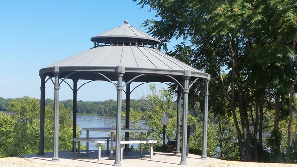 Um gazebo com mesa e bancos, cercado por árvores e um corpo d'água.