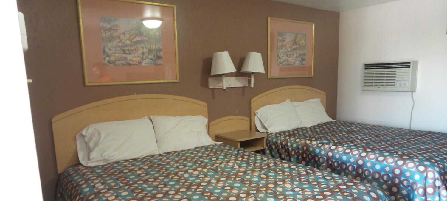 Quarto de hotel com duas camas, cada uma com roupa de cama estampada, mesa de cabeceira, luminárias na parede e duas imagens emolduradas.