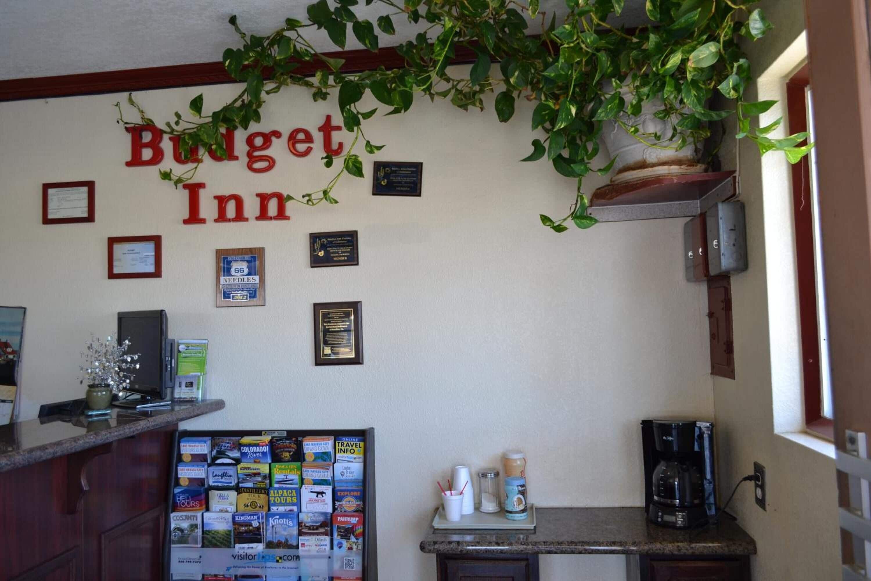 Un área de recepción con un letrero que dice 'Budget Inn', premios enmarcados, un tablón de anuncios con folletos de viajes y una estación de café.