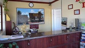 Zona de recepción de hotel con un reloj, un cuadro enmarco de un lugar histórico, un cartel con indicaciones de salida y una planta.