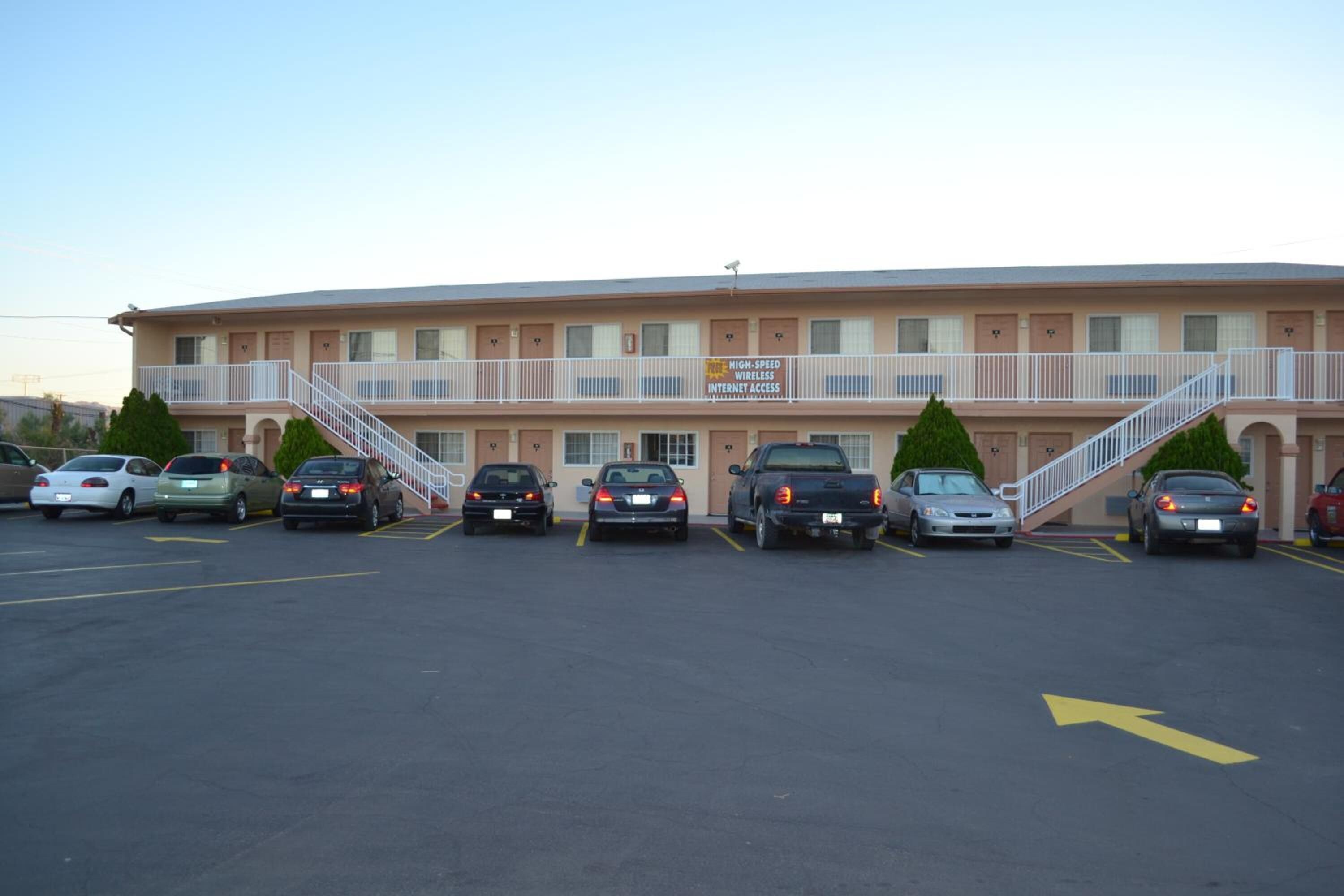 A motel with a sign indicating high-speed wireless internet access. It features a parking lot with several cars parked, a central staircase leading to the entrance, and a flat-roofed building with a beige exterior.