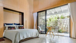 Un dormitorio moderno con una cama, una pequeña mesa con sillas y un ventanal corredero que da a un balcón con plantas.