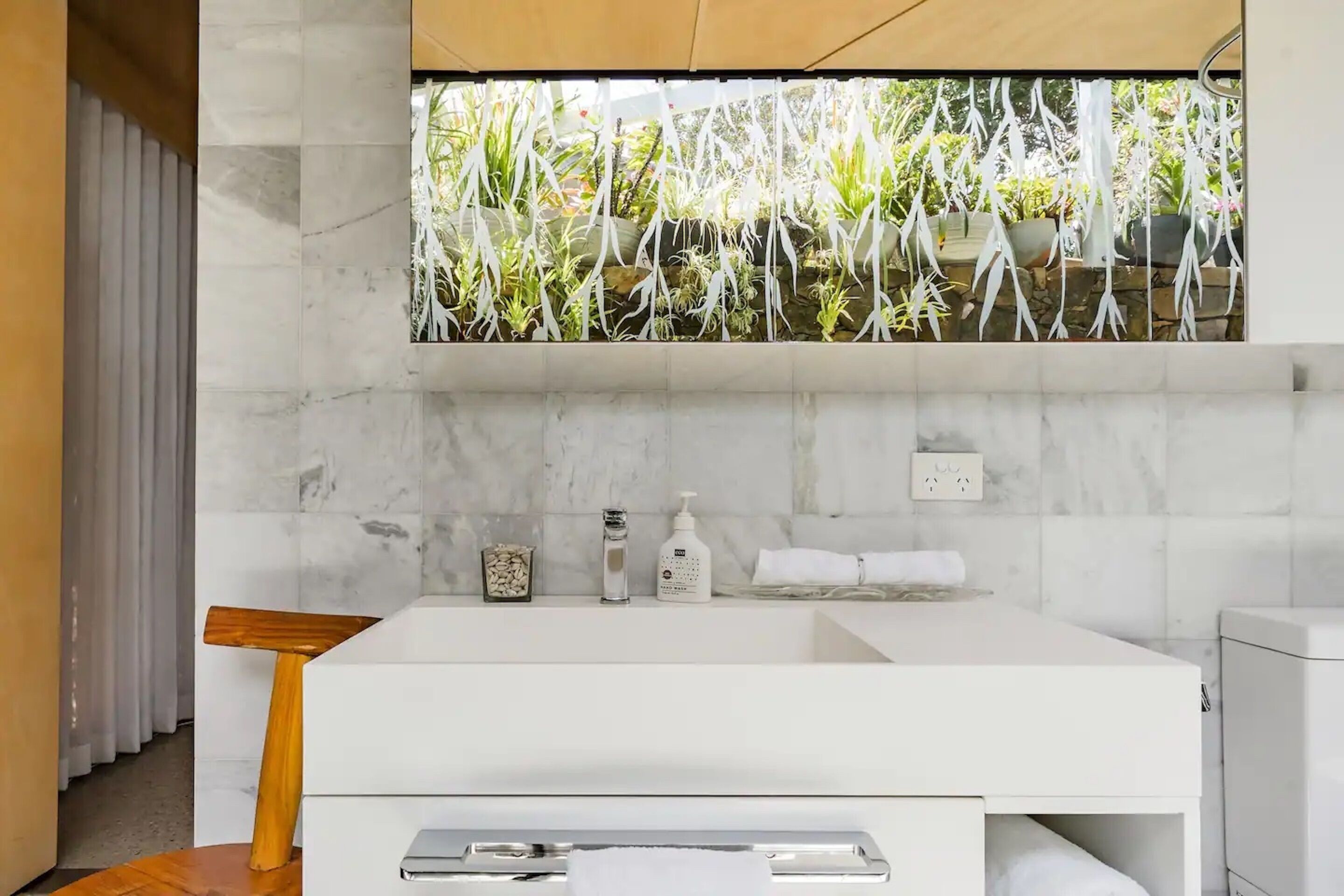 A modern bathroom with a white sink, marble backsplash, and a view of greenery through a window.