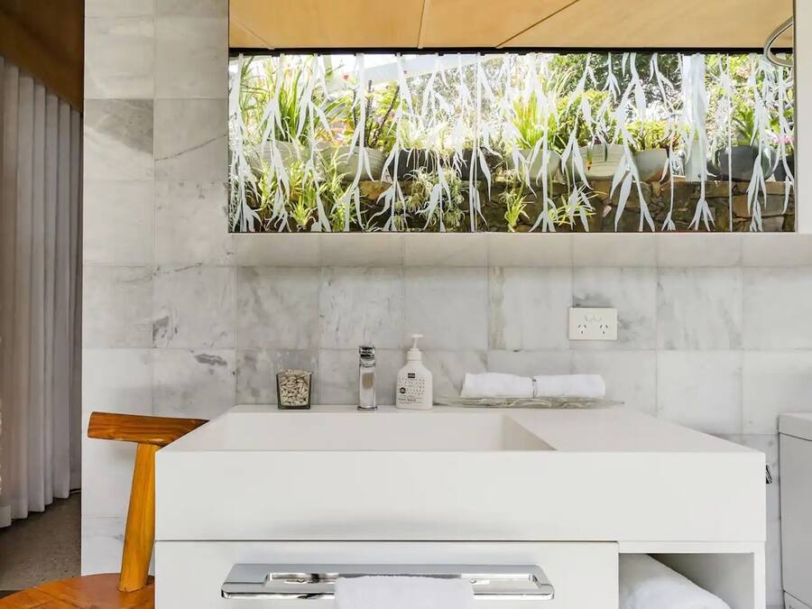 A modern bathroom with a white sink, marble backsplash, and a view of greenery through a window.