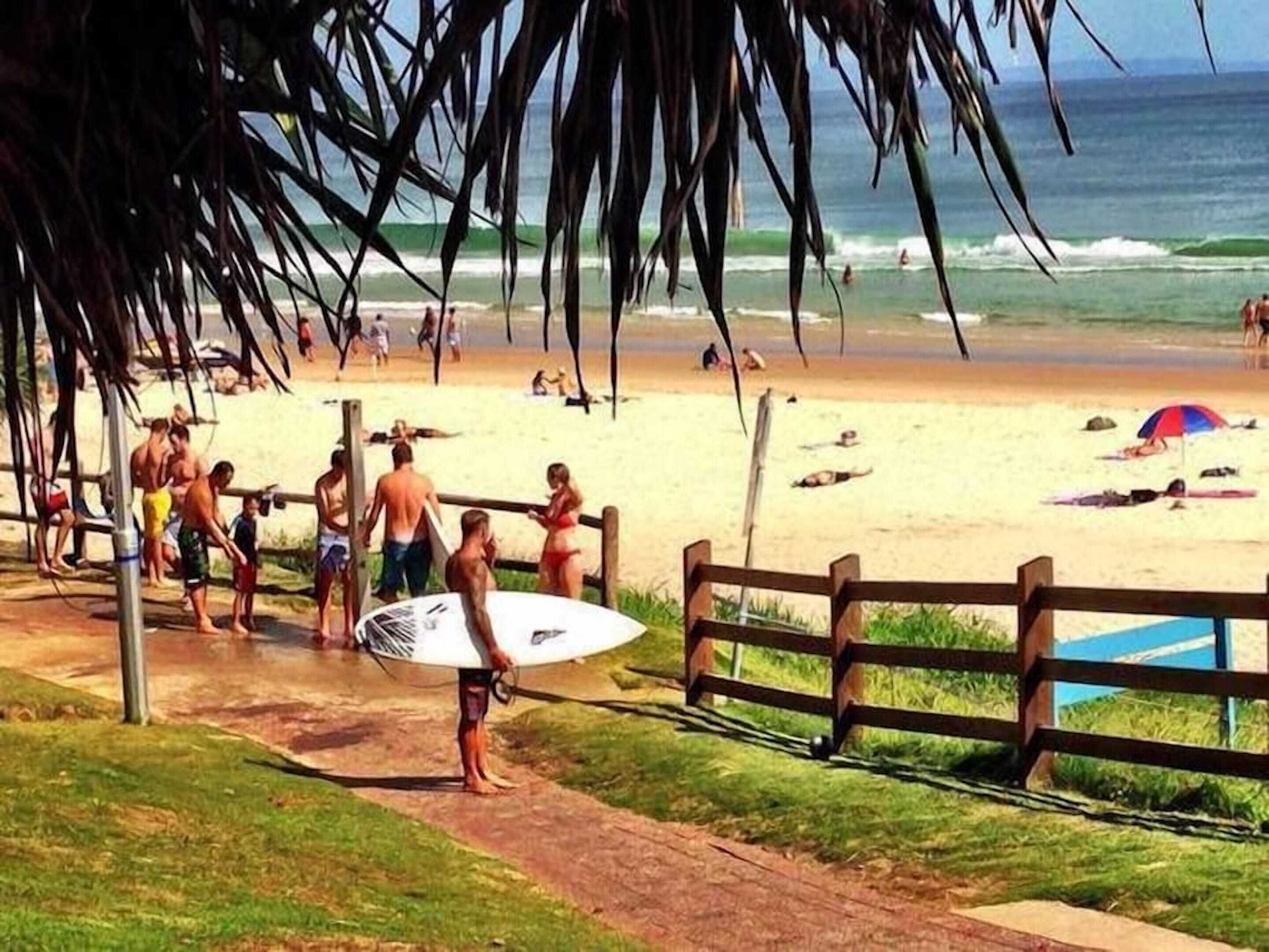 A beach scene with a wooden fence, people walking, and a surfboard.