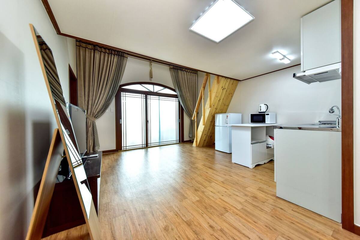 A spacious room with a wooden floor, a kitchenette with a microwave, and a large sliding glass door leading to a balcony.