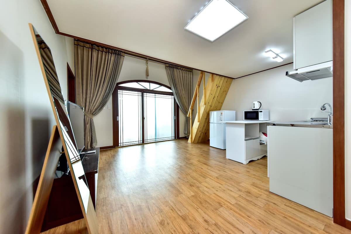 A spacious room with wooden flooring, a large window with curtains, a kitchenette with a microwave, and a ceiling light.