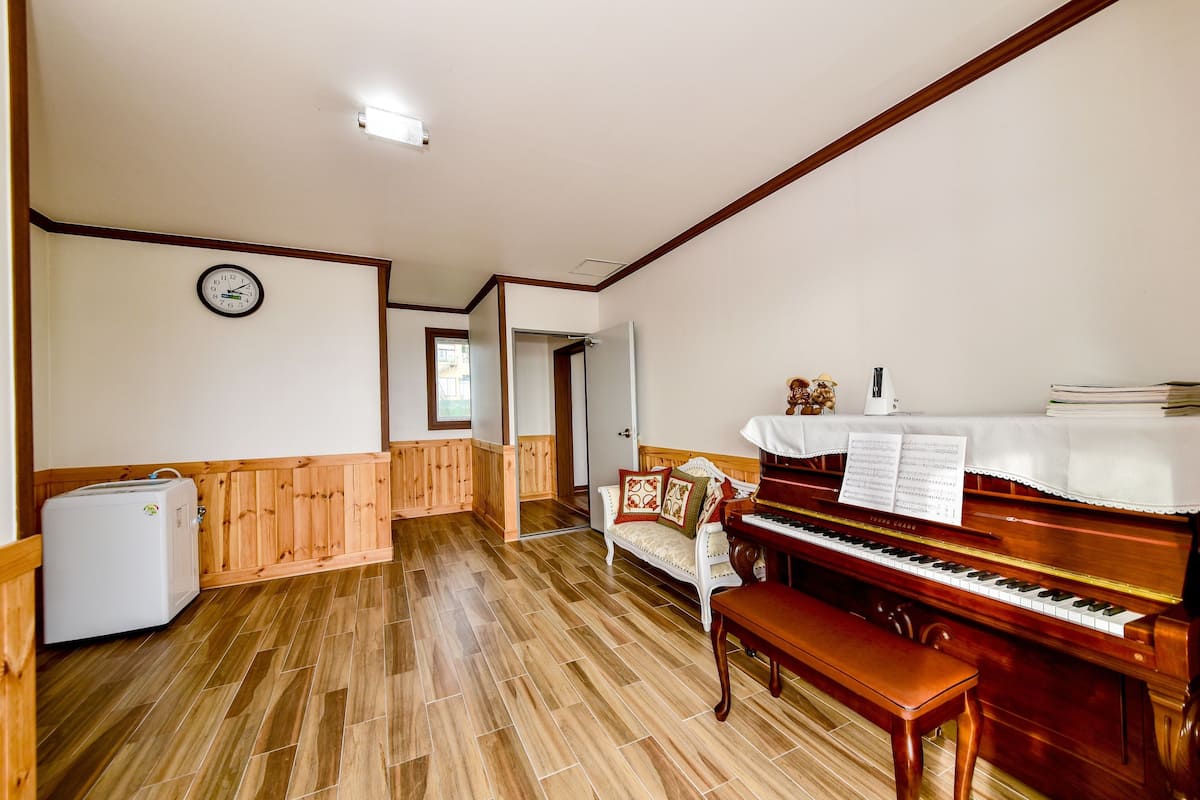 A room with a wooden floor, a piano, a bench, a refrigerator, and a clock on the wall.