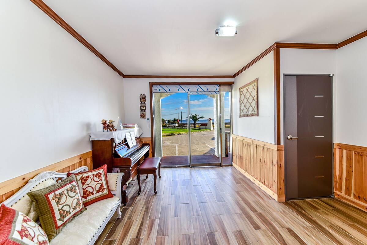 A room with a piano, a bench, a sofa with patterned cushions, and a door leading outside.