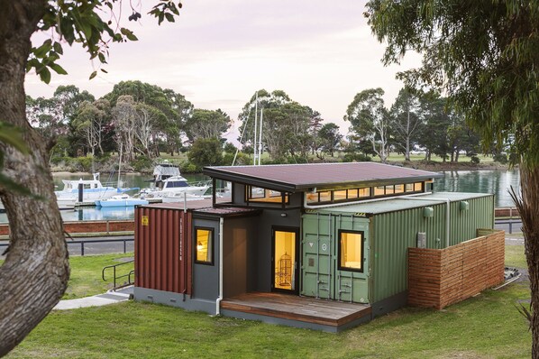 Il s'agit d'une maison moderne et compacte, dotée d'un toit plat et située au bord d'un plan d'eau. Elle comprend une petite terrasse et une porte donnant accès à un espace intérieur.