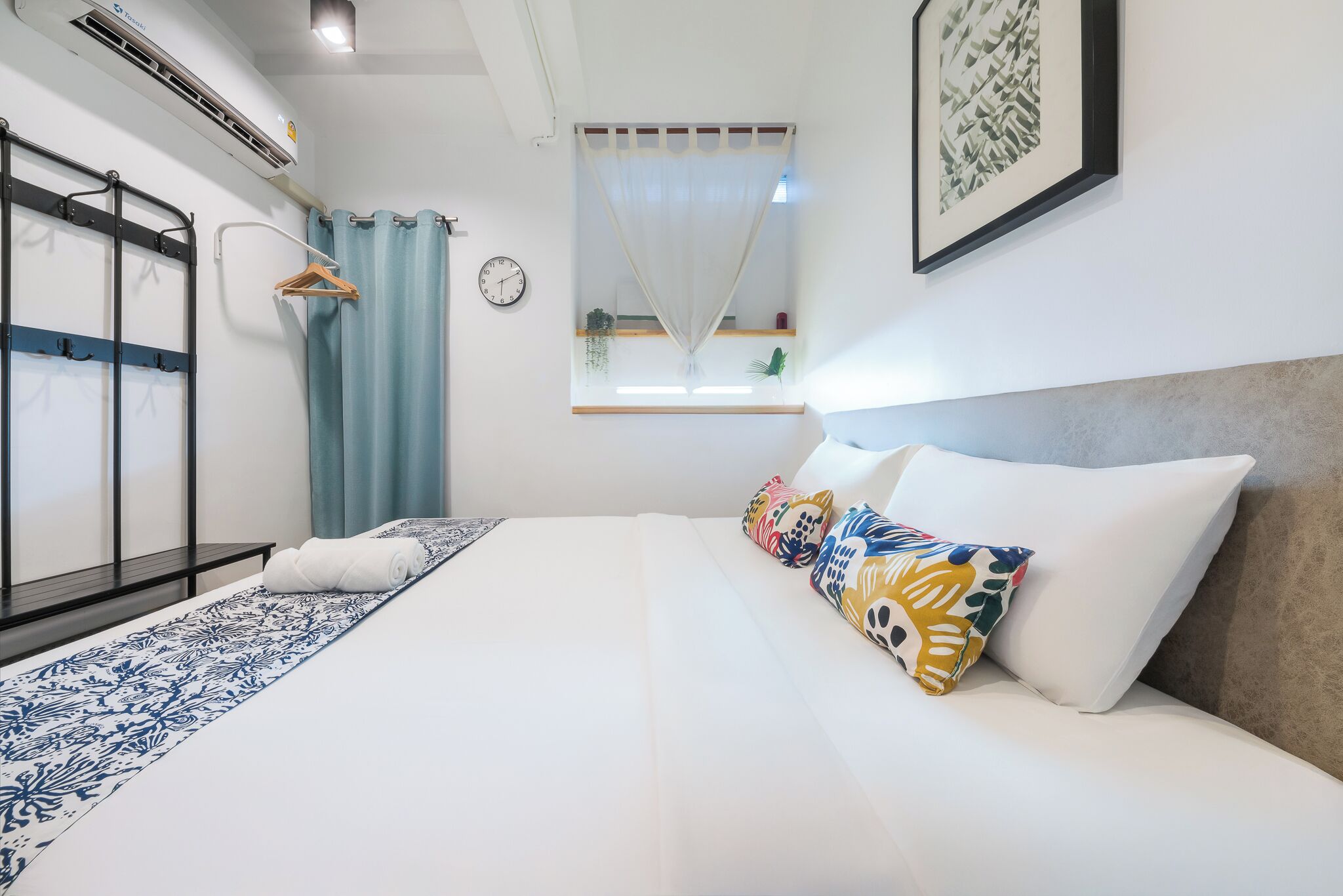 A hotel room with a bed, a window with curtains, a wall clock, and a framed picture.