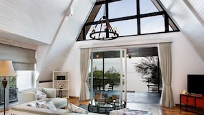 Une chambre spacieuse aménagée en mezzanine, avec un plafond haut, une grande fenêtre, un coin salon confortable et une vue sur l’extérieur.