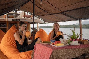 Zwei Personen sitzen auf orangefarbenen Sitzsäcken unter einem Reetdach, vor ihnen steht ein Tisch mit Tellern voller Essen und einer kleinen Pflanze.