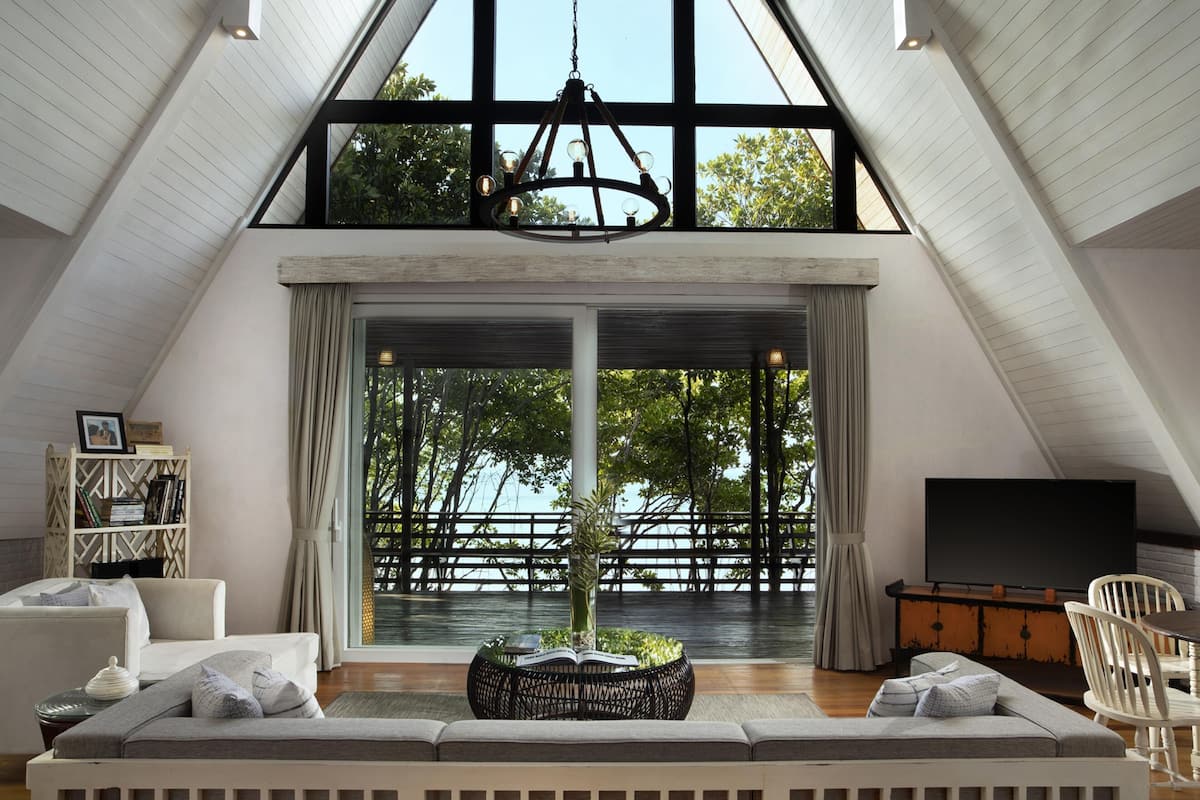 Un loft spacieux avec un plafond haut, une grande fenêtre donnant sur un plan d’eau, un coin salon confortable avec un canapé à motifs et un téléviseur posé sur un meuble en bois.