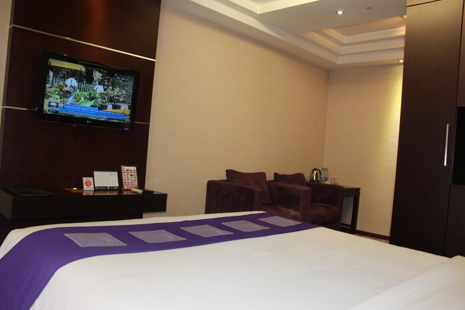 A hotel room with a large bed, a television mounted on the wall, a desk with a calendar, and a seating area with chairs.
