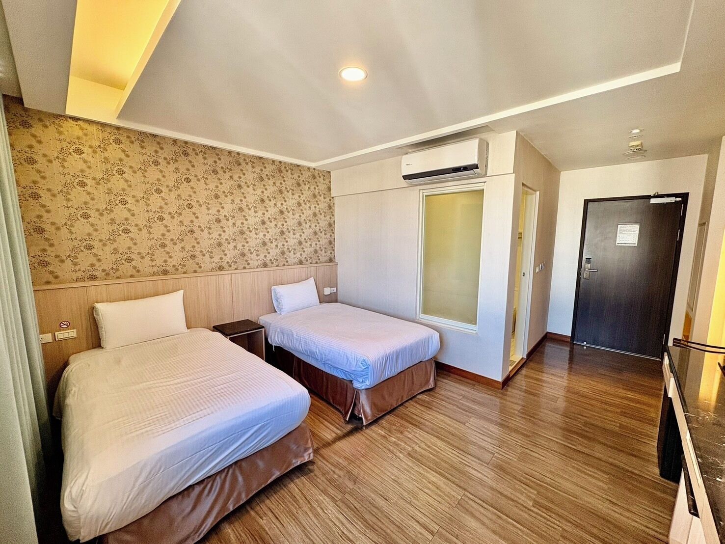 A hotel room with two beds, a wardrobe, an air conditioning unit, and a door.