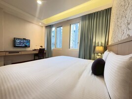 Habitación de hotel con una cama grande, un escritorio con computadora, un televisor en la pared y una ventana con cortinas.
