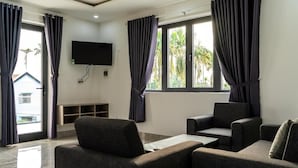 A modern living room with dark grey sofas, a flat-screen TV mounted on the wall, and large windows with curtains. - Rice Field Homestay (Da Nang)