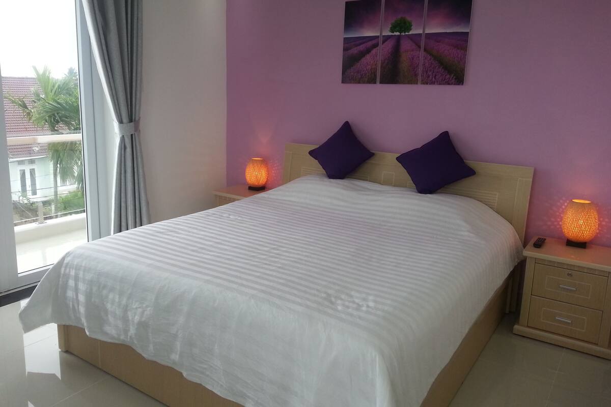 A bedroom with a large bed, two bedside tables with lamps, a window with curtains, and a purple wall with a landscape painting.