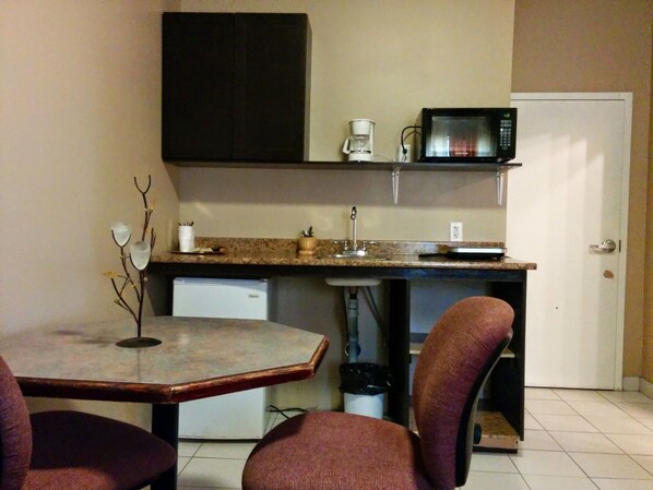 Cocina compacta con microondas, cafetera y un refrigerador pequeño. Hay una mesa de comedor con dos sillas y una planta decorativa.