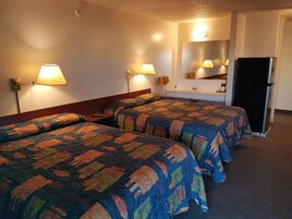 A hotel room with two beds, a dresser, a mirror, a refrigerator, and wall-mounted lamps.