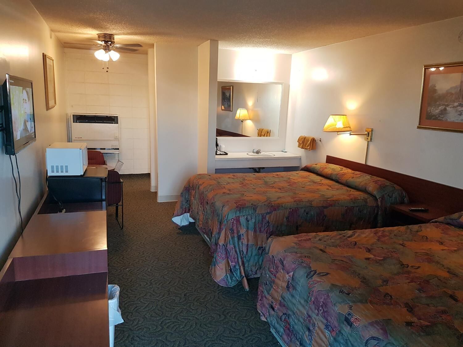 A hotel room with two beds, a desk, a chair, a TV, a ceiling fan, a mirror, and a window.