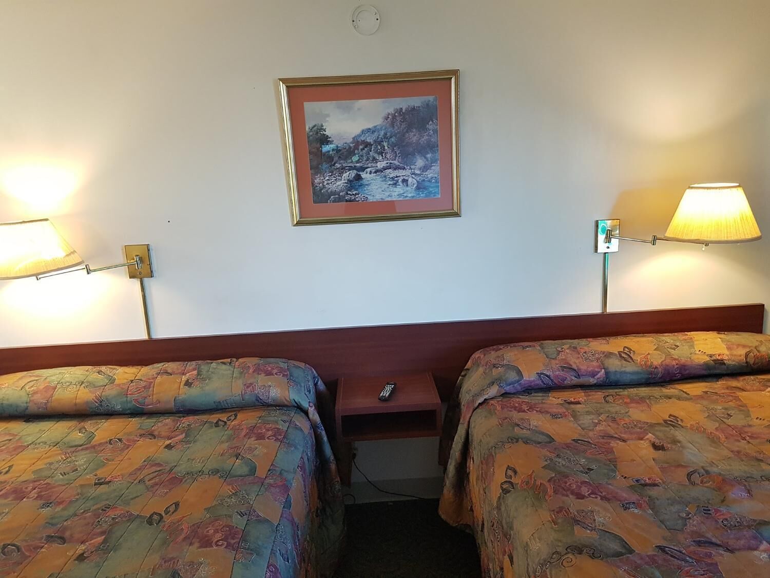 Quarto de hotel com duas camas, um quadro na parede e duas luminárias fixadas na parede.