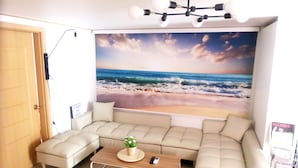A living room with a beige sectional sofa, a wooden coffee table with a plant, a wall-mounted light fixture, and a beach mural. - Hongdae Lai Lai house (Seoul)