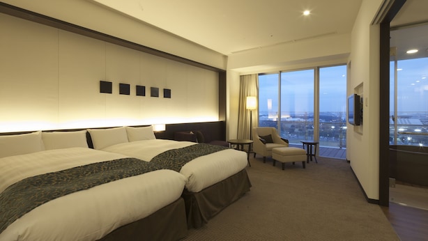 Habitación de hotel con dos camas, una mesita, una silla y un amplio ventanal con vistas a la ciudad.