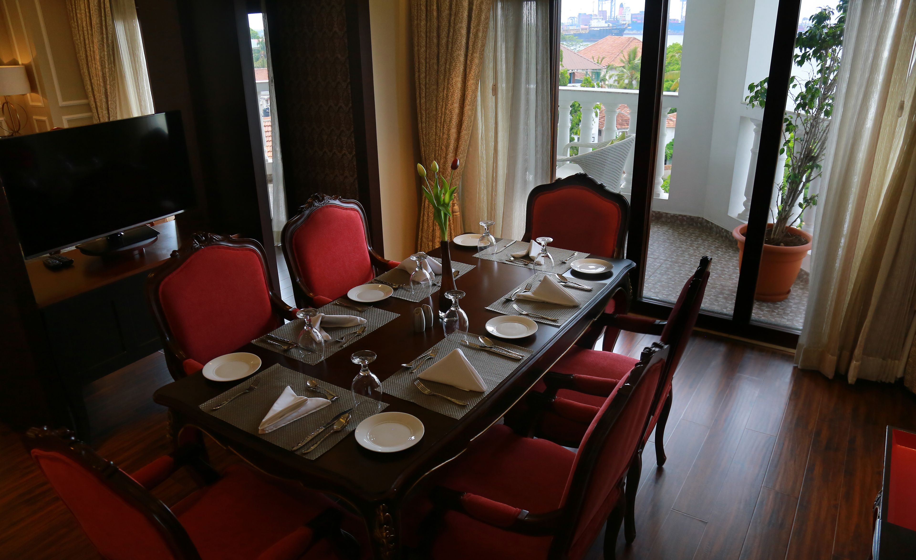 A dining area with a long table set for six, red upholstered chairs, a vase with a plant, and a television on the left.